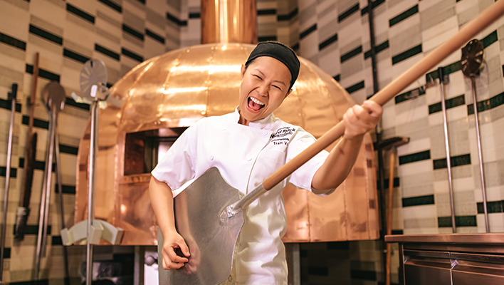 Woman standing in front of pizza oven pretending to play an air guitar